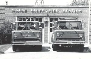 Two vintage Ford fire trucks are parked in front of the Maple Bluff Fire Station, a brick building with large double doors and signage above the entrance. The image is in black and white.