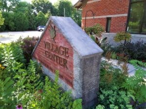 A brick and stone sign reading Village Center, surrounded by lush greenery and flowers, stands near a red-brick building on a sunny day.