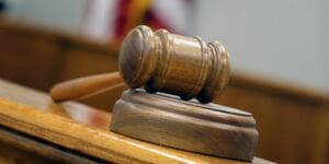 A close-up view of a wooden judges gavel resting on a sound block in a courtroom setting, with blurred background elements.