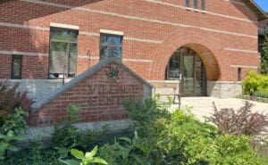 A red brick building with arched windows and doorways labeled Village Center on a stone sign surrounded by green plants and flowers in front.