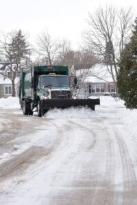 A green snow plow truck clears snow from a residential street lined with snow-covered houses and trees on a winter day.