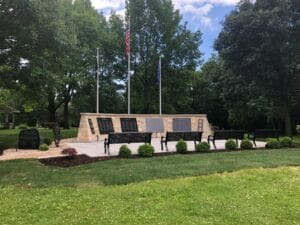 A stone memorial wall with plaques stands in a park, flanked by three flagpoles and black metal benches. Green grass, shrubs, and large trees surround the area under a partly cloudy sky.
