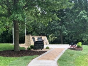 A stone monument with plaques stands beside a path surrounded by green grass, trees, benches, and landscaping; the sign reads Mary Blair Veterans Memorial.