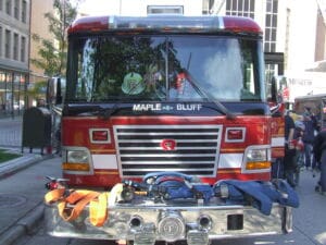 A red Maple Bluff fire truck is parked on a city street, with firefighting hoses and equipment displayed on its front bumper. People and buildings are visible in the background.