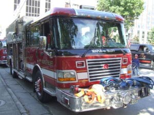 A red Maple Bluff fire truck is parked on a city street on a sunny day. Coiled hoses are attached to the front bumper, and people, including a child, are visible nearby on the sidewalk.