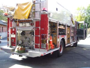 A red fire truck is parked outdoors with its side and back compartments open, showing hoses, tools, and equipment. The truck has a white roof, an open door, and sunlight illuminating the scene.