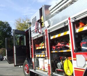 A close-up of a fire truck with its side compartments open, displaying various firefighting equipment and gear, such as hoses, bags, and tools, under clear skies in a parking lot.