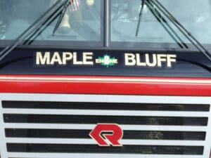 Close-up of the front of a vehicle with the words MAPLE BLUFF ENGINE in gold letters and a red emblem below a red horizontal stripe. The windshield and reflections are visible at the top.
