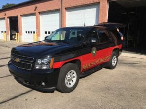 A black and red Maple Bluff emergency SUV is parked outside a brick building with several garage doors. The vehicles back hatch is open, and a person is partially visible in the background.
