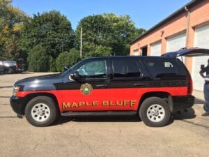A black and red SUV marked Maple Bluff Fire Rescue is parked outside a brick building with multiple garage doors on a sunny day.