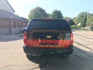 A black SUV with MAPLE BLUFF written in large yellow letters on the back, parked on a sunny street near brick buildings and trees.