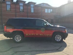 A black and red Maple Bluff fire rescue SUV labeled CAR 1 is parked outside a brick building on a sunny day.