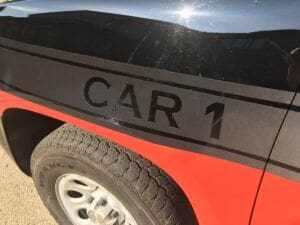 Close-up of the side of a black and red vehicle with the text CAR 1 in large black letters above the front wheel. The tire brand is visible, and reflections can be seen on the cars surface.