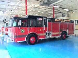 A red fire truck labeled Maple Bluff Fire Rescue is parked inside a brightly lit fire station with a blue floor and several other emergency vehicles in the background.