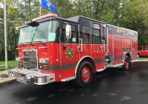 A red Maple Bluff fire engine is parked on wet pavement near green trees, with a blue flag visible in the background. The vehicle displays firefighting insignia and has shiny chrome details.