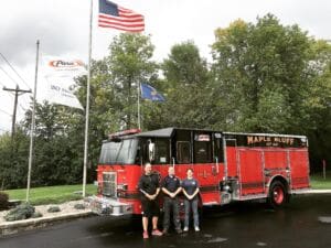 Three people stand in front of a red Maple Bluff fire truck parked on wet pavement, with flags flying on tall poles and green trees in the background under a cloudy sky.