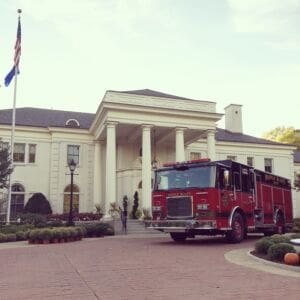 A red fire truck labeled Maple Bluff is parked on a brick driveway in front of a large white mansion with tall columns. Potted plants and an American flag are visible near the entrance.