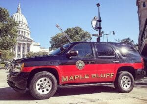 A black and red Maple Bluff fire department SUV is parked on a city street, with a domed government building and trees visible in the background under a clear blue sky.