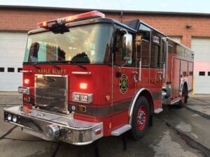 A bright red Maple Bluff fire truck is parked in front of a building with white garage doors, reflecting the sky and surroundings on its shiny surface.