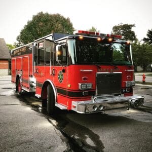 A red Maple Bluff fire truck is parked on a street near trees and buildings, reflecting light on its shiny chrome bumper and trim.