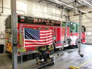 A red fire truck labeled MAPLE BLUFF EST 1947 is parked inside a fire station. An American flag is draped over a metal rack with firefighting gear beneath it. Large hoses hang from the ceiling.