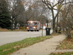 A fire truck with flashing lights drives down a suburban street lined with trees and houses; fallen leaves are scattered along the sidewalk and grass.