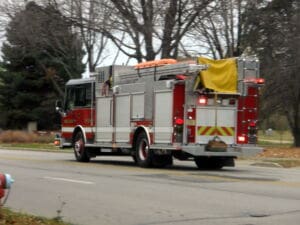 A red and white fire truck with lights on drives down a suburban street lined with bare trees on a cloudy day.