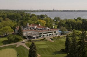 Aerial view of a large clubhouse with green lawns, trees, and a golf course near a lake, with a town and distant buildings visible across the water under a clear sky.