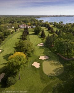 Aerial view of a lush green golf course with trees, sand bunkers, and putting greens, set near a large lake and surrounded by dense foliage and distant buildings.