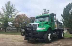 A large green and silver dump truck is parked on a paved area near trees and houses on a cloudy day.