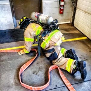 A firefighter in full gear crawls on hands and knees along a concrete floor, holding a fire hose, with a breathing apparatus on their back inside a fire station.
