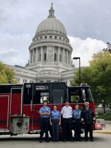 Five firefighters stand in front of a red fire truck, with a large white-domed government building in the background, surrounded by green trees. The group poses for the photo, smiling, in uniform.