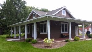 A small brown house with white trim and a wraparound porch sits on a manicured lawn, surrounded by shrubs and trees. A wooden bench and a trash can are visible near the front entrance.