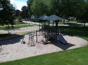 A playground with climbing structures, slides, and swings sits on mulch surrounded by green grass and trees, with nearby houses and a street visible in the background on a sunny day.
