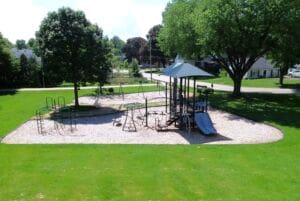 A playground with slides, climbing structures, and swings sits on a bed of gravel, surrounded by green grass and trees in a suburban neighborhood. Houses and a street are visible in the background.