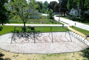 A playground with empty swings is set on wood chips in a residential neighborhood, surrounded by green grass, trees, and houses on a sunny day. A row of bike racks lines one edge of the playground.