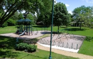 A playground in a park with slides, climbing structures, swings, bike racks, and trees providing shade under a blue sky. A sign reads Freemens Park, and a path leads through the grassy area.