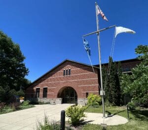 A brick building with arched entrance sits under a clear blue sky. Three flags are hoisted on a pole in front, and trees line the sidewalk and landscaped greenery.