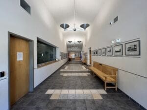A bright, empty hallway with tiled floors, a wooden bench on the right, framed photos on the wall, a glass window to the left, and overhead modern light fixtures. A clock is visible at the far end.