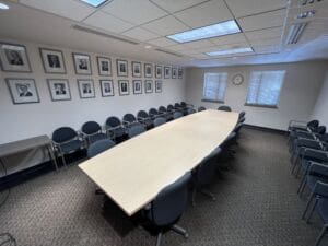 A large conference room with an oval table surrounded by chairs. The walls are adorned with framed black-and-white portraits, and two windows and a clock are visible at the far end of the room.