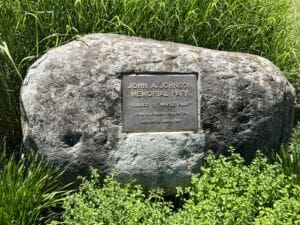 A large rock with a plaque reading John A. Johnson Memorial Park, Village of Maple Bluff, John A. Johnson Foundation, Dedicated 1976, surrounded by grass and greenery.