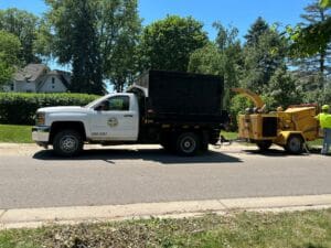 A white municipal truck is parked on the street next to a yellow wood chipper. Two workers in neon safety gear stand nearby, surrounded by trees and residential homes on a sunny day.