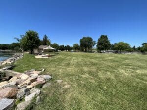 A large grassy park area beside a lake, bordered by rocks. Trees and a small building with a tent canopy are visible in the background under a clear blue sky.