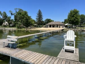 A wooden dock extends over greenish water toward a pavilion on shore, with white benches and a chair on the dock, surrounded by trees and a clear blue sky.