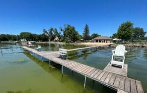 A wooden dock with benches and a white chair extends over clear, shallow water toward a sandy beach and a small building, surrounded by green trees under a bright blue sky.
