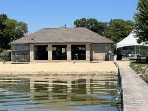 A small building with a brown roof and tan brick walls sits on a sandy beach by a calm lake, with a wooden dock leading up to it. Trees and a white tent are visible in the background under a clear blue sky.