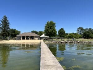 A wooden dock extends over clear, greenish water toward a sandy shore with a small brick building and trees under a bright blue sky. People are visible near the rocks along the shoreline.