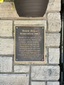 A bronze plaque on a stone wall reads: “Beach House Renovation 2003. Thanks to the generosity of Don and Dolly Warren and family… to restore this beloved facility and landmark.” A flower pot hangs above.