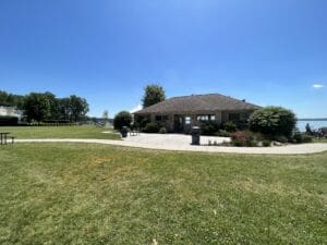 A small building with bushes and flowers around it sits in a park with a grassy lawn, paved walkways, and trees. The sky is clear and blue, and a body of water is visible in the background.