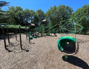 A playground with climbing structures, monkey bars, slides, and a geometric climbing dome sits on a mulch surface, surrounded by trees under a clear blue sky.